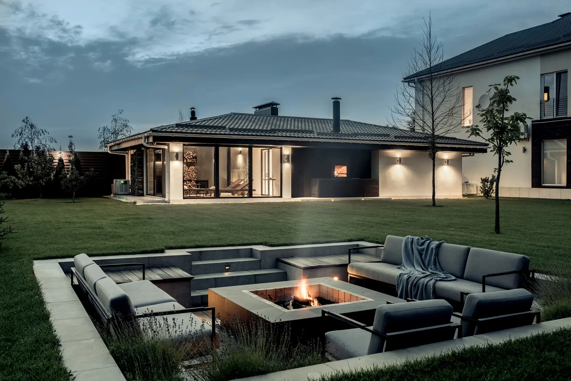 Sunken paved fire-pit lounge with built-in bench seating and steps up to a large lawn at dusk
