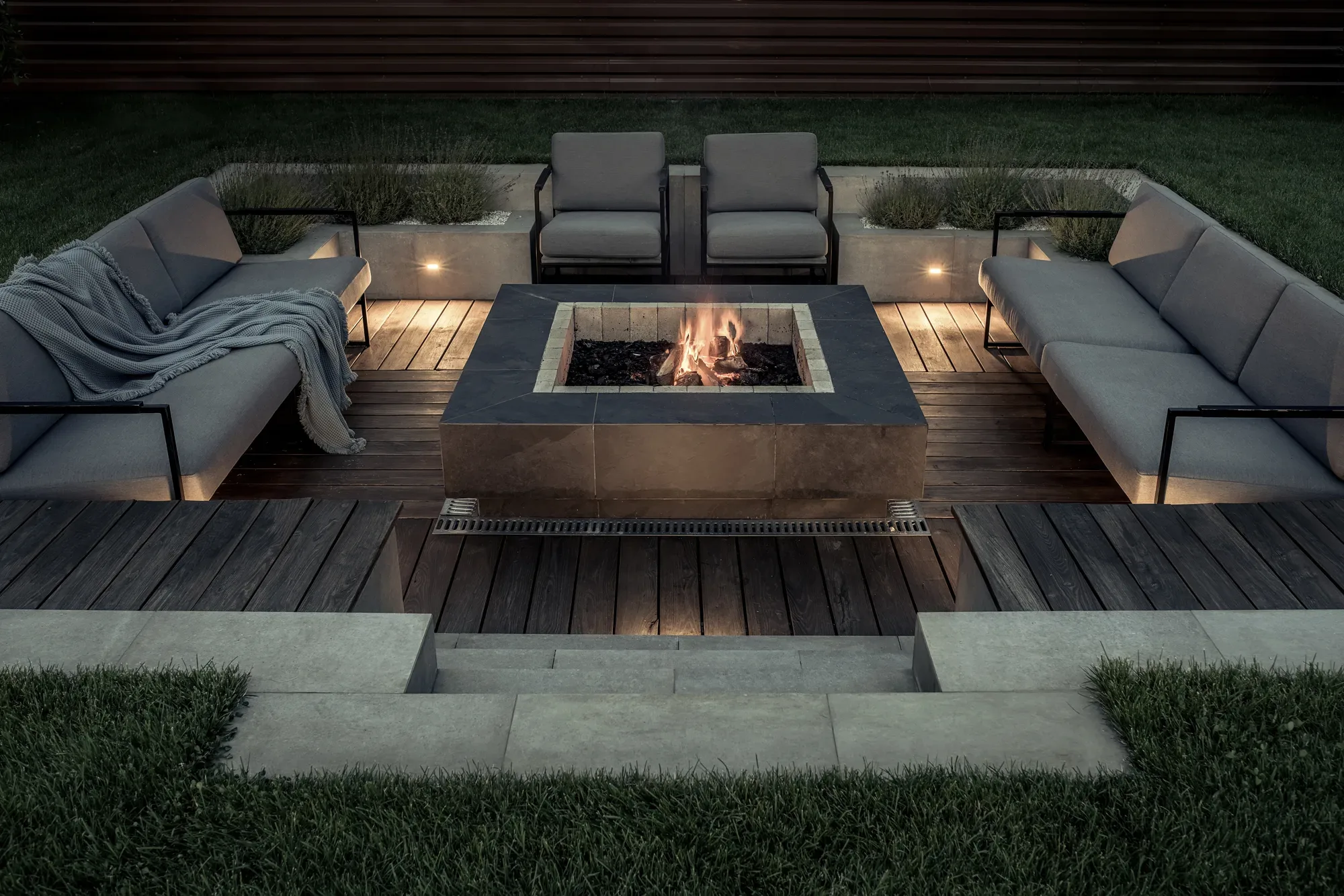 Sunken fire-pit lounge with a stone-clad fire table, grey sectional sofas and timber deck at night