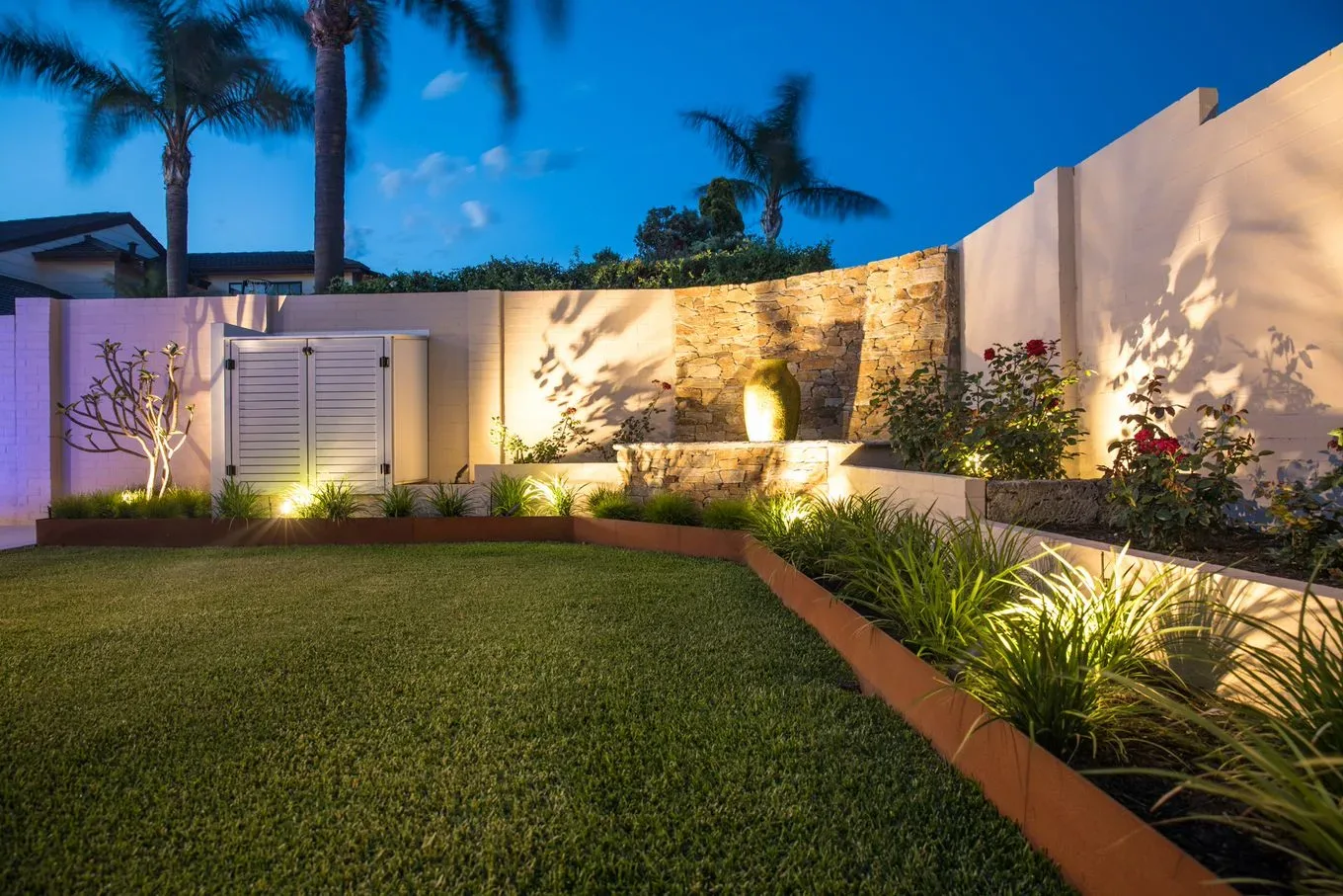 Curved rendered retaining wall with a stacked-stone water feature insert and garden lighting at night